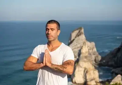 Ein Mann steht mit gefalteten Händen auf einem Felsen und atmet tief durch, im Hintergrund sieht man das Meer